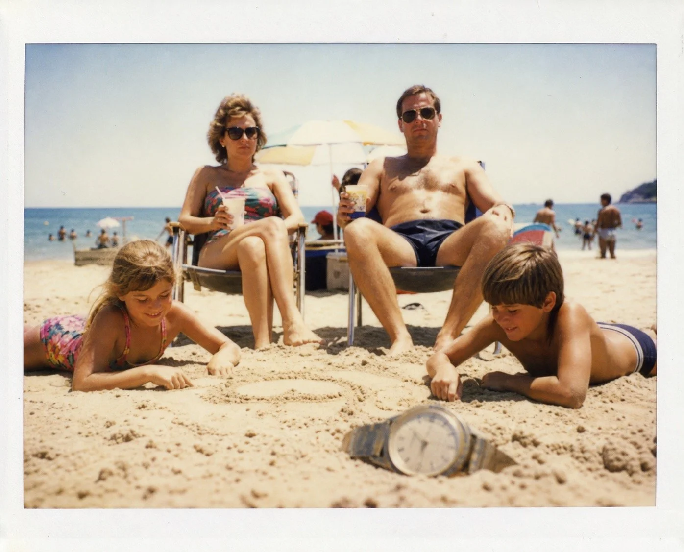 A faded holiday photograph of a family on a beach beside a wristwatch buried in the sand